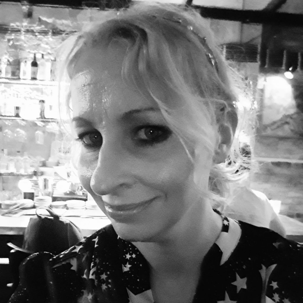 Heather is smiling at the camera. She is standing in front of a public bar, and is wearing a stylish black and white blouse with a star pattern.