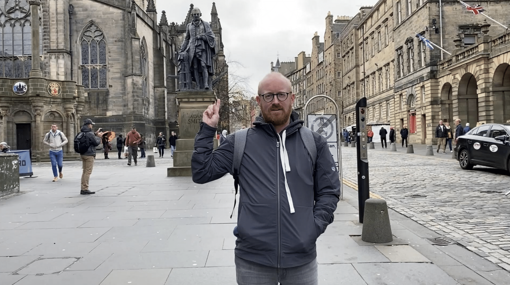 Matt May standing on the Royal Mile in Edinburgh points at the Adam Smith statue.