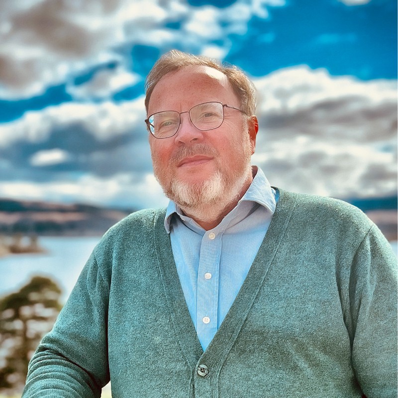 Alistair is shown outside against a bright blue sky and water. His hair is in a side parting, and he has a short white beard and thin-rimmed glasses. He is wearing a light blue shirt and light grey cardigan.