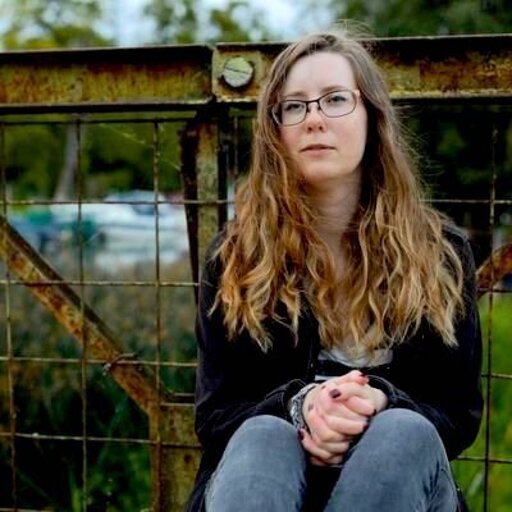 Beverly is wearing dark-rimmed glasses; they have long, fair hair blowing in the breeze. They are outdoors, squatting in front of a metal gate.